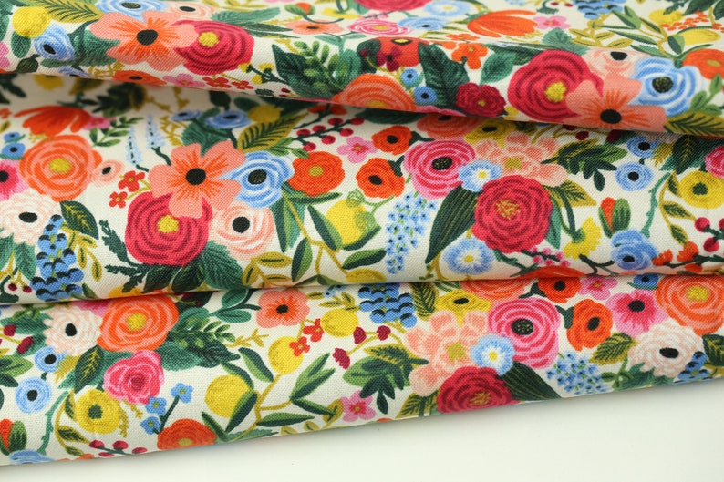 May include: Folded fabric with a colourful floral design. The pattern showcases red, pink, orange, blue, and yellow flowers on a cream backdrop. Green leaves and stems are also visible, creating a botanical theme.