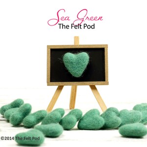 Puede incluir: Corazones de fieltro verde mar sobre una superficie de madera blanca. Los corazones est&aacute;n dispuestos en una fila, con un coraz&oacute;n m&aacute;s grande en un caballete de pizarra en el fondo. El caballete de pizarra tiene el texto "Sea Green The Felt Pod" escrito en &eacute;l.