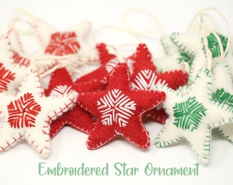 Puede incluir: Un grupo de adornos de estrellas de fieltro con copos de nieve bordados. Las estrellas son de color rojo, blanco y verde.