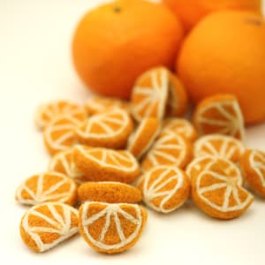 May include: A group of felt orange slices in various sizes, with a few whole oranges in the background. The felt orange slices are a light orange color with white stitching to create the segments.