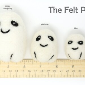 May include: Three white felt pod toys with black embroidered eyes and smiling mouths. The toys are lined up from largest to smallest, with a ruler underneath to show their size. The largest pod is labeled "Large (original)", the middle pod is labeled "Medium", and the smallest pod is labeled "Mini". The text "The Felt Pod" is at the top of the image.