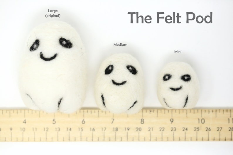 May include: Three white felt pod toys with black embroidered eyes and smiling mouths. The toys are lined up from largest to smallest, with a ruler underneath to show their size. The largest pod is labeled "Large (original)", the middle pod is labeled "Medium", and the smallest pod is labeled "Mini". The text "The Felt Pod" is at the top of the image.