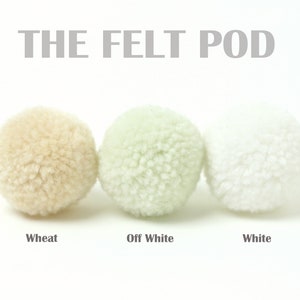 Pu&ograve; includere: Tre palline di feltro in diverse tonalit&agrave; di bianco: grano, bianco avorio e bianco. Le palline di feltro sono disposte in fila da sinistra a destra. Il testo "THE FELT POD" &egrave; sopra le palline.