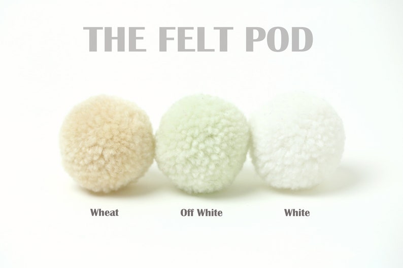Pu&ograve; includere: Tre palline di feltro in diverse tonalit&agrave; di bianco: grano, bianco avorio e bianco. Le palline di feltro sono disposte in fila da sinistra a destra. Il testo "THE FELT POD" &egrave; sopra le palline.