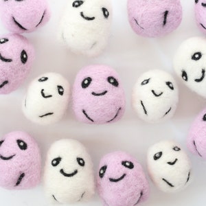 May include: A collection of 14 felt ghost ornaments in pink and white with various facial expressions. The ghosts are all approximately the same size and are arranged in a random pattern on a white background.