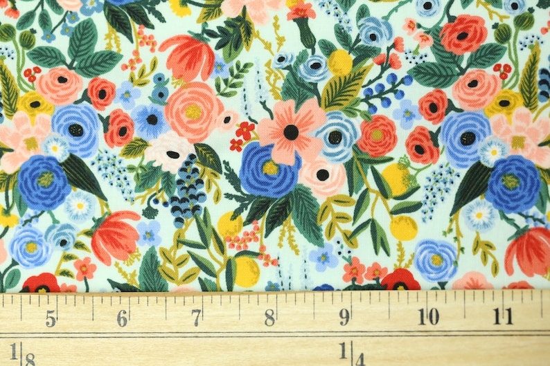 May include: Floral fabric with a pale blue backdrop. The fabric is densely patterned with flowers in red, pink, blue, yellow, and green. A wooden ruler with inch markings is at the bottom.