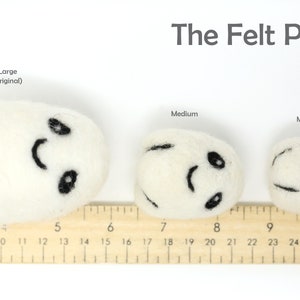May include: Three felt pods of varying sizes, labeled "Large (original)", "Medium", and "Mini". Each pod is white with a black smiling face and is measured against a wooden ruler.