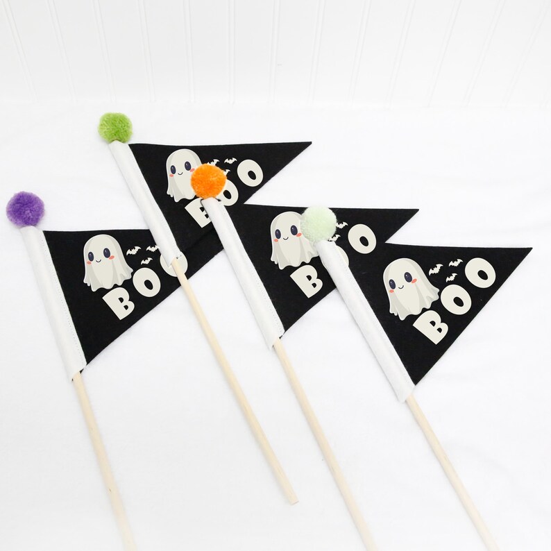 Op de afbeelding: Drie zwarte driehoekige vlaggen met een witte rand en een schattig spookontwerp. Elke vlag heeft het woord "BOO" erop gedrukt in witte letters. De vlaggen zijn bevestigd aan houten stokken met kleurrijke pompoenen aan de bovenkant.