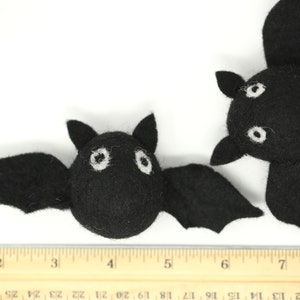May include: Two black felt bats with white eyes and wings are displayed against a white background. A wooden ruler is placed below the bats. The bats are handmade and likely for Halloween decorations.