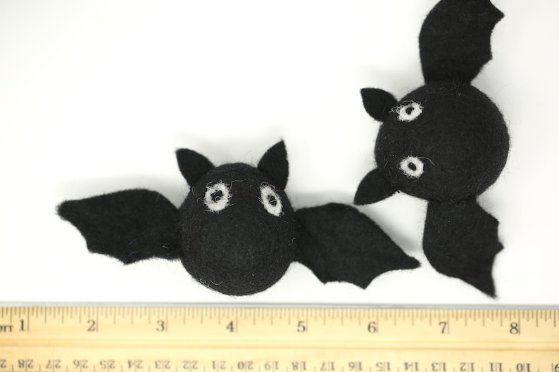 May include: Two black felt bats with white eyes and wings are displayed against a white background. A wooden ruler is placed below the bats. The bats are handmade and likely for Halloween decorations.