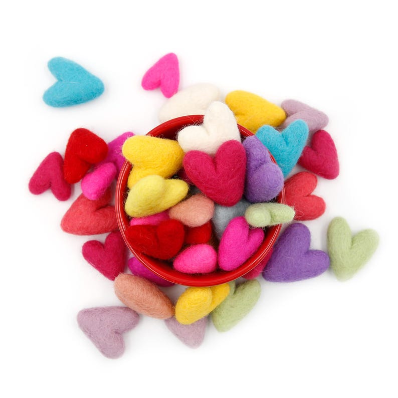 May include: A red bowl filled with felt hearts in various colors, including pink, red, blue, green, yellow, and white. The hearts are scattered around the bowl on a white background.