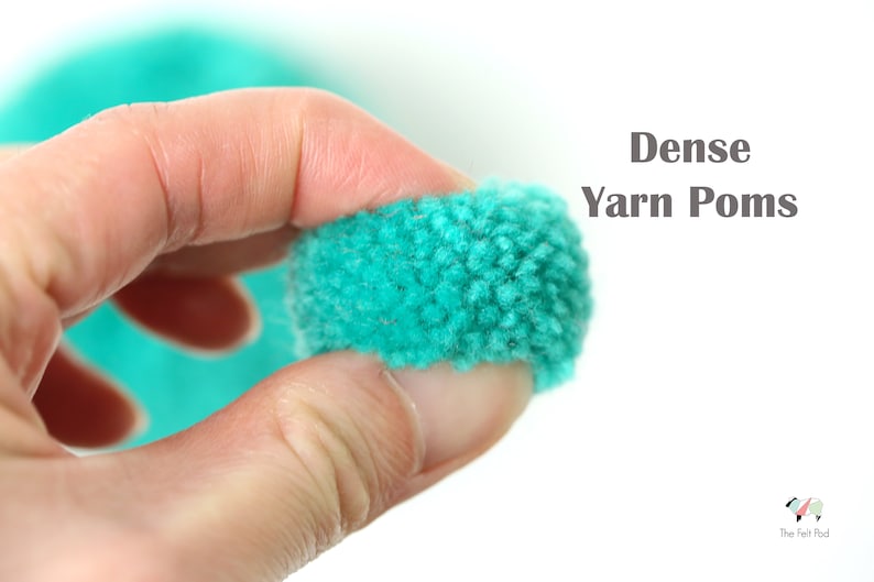 Pu&ograve; includere: Una mano tiene un pompon di lana color turchese brillante. Il pompon &egrave; molto denso e soffice. Il testo "Dense Yarn Pom Poms" &egrave; visibile nell'immagine.