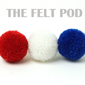 May include: Three felt balls in red, white, and blue colours. The felt balls are arranged in a row from left to right. The text "THE FELT POD" is above the balls.