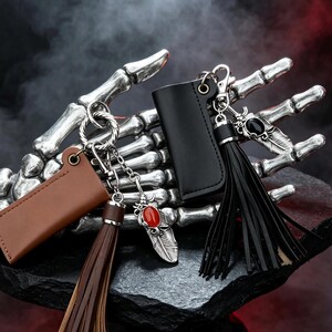 Könnte beinhalten: Zwei Schlüsseletuis aus Leder mit passenden Quasten und Metallfederanhängern. Ein Etui ist braun mit einem roten Stein, das andere schwarz mit einem schwarzen Stein. Beide verfügen über silberfarbene Beschläge, Karabinerhaken und Ringe.