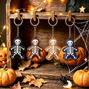 May include: Four keychains shaped like gingerbread people with white skeleton prints. Each keychain features a metal ring and chain, with one black, one dark brown, one light brown, and one navy blue leather-like material skeleton figure.