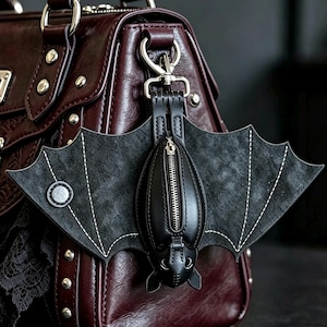 Peut inclure: Un porte-monnaie noir en forme de chauve-souris avec une fermeture éclair argentée centrale, des ailes texturées à surpiqûres blanches et un bouton-pression circulaire. L'accessoire est fixé à un sac à main bordeaux par un mousqueton argenté.