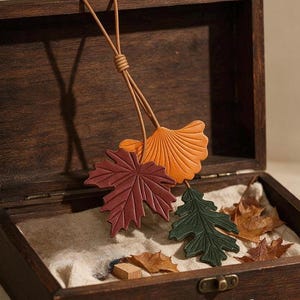 Può includere: Un ornamento sospeso composto da tre foglie in pelle color arancione, rosso scuro e verde scuro, appese a un sottile cordino di pelle color cuoio. Le foglie riproducono le forme di ginkgo, acero e quercia con venature e texture in rilievo.