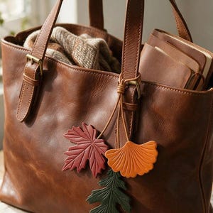 Könnte beinhalten: Eine braune Ledertasche mit zwei Tragegriffen und messingfarbenen Schnallen. An einem Griff befindet sich ein dekorativer Anhänger aus drei verschiedenen Lederblattformen in Rot, Orange und Dunkelgrün, die an dünnen Lederbändern befestigt sind.