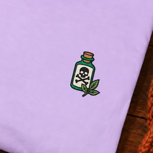 May include: A light purple crewneck t-shirt featuring a small embroidered graphic on the left chest. The graphic depicts a green glass bottle with a cork stopper, a white skull and crossbones symbol, and two green leaves at the base.