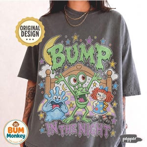 May include: A charcoal gray t-shirt featuring a graphic print with the text BUMP IN THE NIGHT. The design includes a green monster, a blue blob creature, and a doll standing in front of a bed, surrounded by stars and a crescent moon.