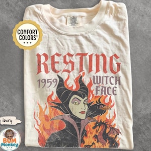 May include: An ivory t-shirt featuring a graphic print of a character with horns, a black bird, and a dragon surrounded by orange and red flames. The text reads RESTING 1959 WITCH FACE. The shirt has a crew neckline and a Comfort Colors brand label.