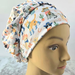 May include: A scrub cap featuring a repeating pattern of cats wearing cowboy hats, bandanas, and boots, interspersed with cacti and horseshoes on a white background with light gray horizontal stripes. The cap has a pleated design at the front.