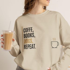 Könnte beinhalten: Ein beigefarbenes Sweatshirt mit Rundhalsausschnitt und dem schwarz-goldenen Text "COFFE, BOOKS, DOGS, REPEAT.", der vertikal auf der linken Seite gedruckt ist, ergänzt durch eine einfache schwarze Strichzeichnung einer dampfenden Kaffeetasse.