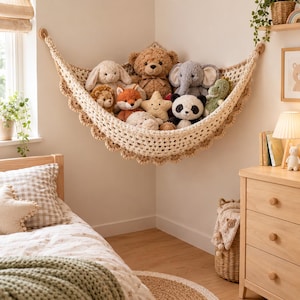 Op de afbeelding: Een crèmekleurige gehaakte hoekhangmat gemonteerd aan de muur, gevuld met diverse knuffels waaronder een bruine teddybeer, een grijze olifant, een panda, een vos, een leeuw, een stervormig kussen en andere diverse knuffeldierfiguren.