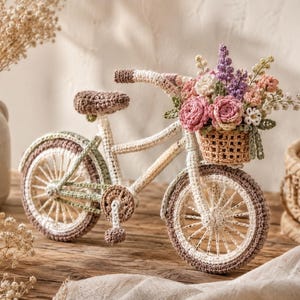May include: A crocheted bicycle figurine in cream, brown, and sage green yarn. The bicycle features a front basket filled with crocheted flowers in shades of pink, purple, and white, including roses and sprigs, with a brown seat and pedals.
