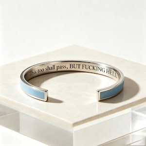 May include: A silver-toned metal cuff bracelet featuring a light blue enamel inlay on the exterior. The interior surface of the bracelet is engraved with the black text, "This too shall pass, BUT FUCKING HELL," in a clear, serif typeface.