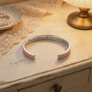 May include: A silver-toned metal cuff bracelet featuring a soft pink enamel inlay along the outer band. The interior surface is engraved with the black text: This too shall pass, BUT FUCKING HELL. The bracelet has an open design for easy wear.
