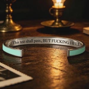 May include: A silver-toned cuff bracelet featuring a light mint green inlay on the exterior and the engraved text "This too shall pass, BUT FUCKING HELL" on the interior surface. The bracelet has an open design with a smooth, polished finish.