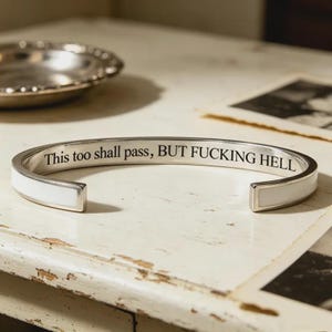 May include: A silver-toned cuff bracelet featuring a white inlay along the outer band. The interior surface of the metal bracelet is engraved with the black text: This too shall pass, BUT FUCKING HELL. The piece has an open design for adjustment.