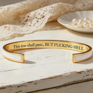 May include: A gold-tone metal cuff bracelet featuring a white enamel inlay along the outer band. The interior surface is engraved with the black text, This too shall pass, BUT FUCKING HELL, in a clear serif font, creating a bold and contrasting look.