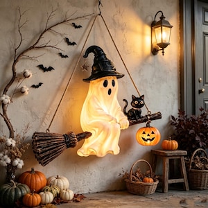 Könnte beinhalten: Eine hängende Halloween-Dekoration mit einem leuchtenden weißen Geist in einem schwarzen Hexenhut, der auf einem Besenstiel sitzt, zusammen mit einer schwarzen Katze und einem kleinen orangefarbenen Kürbis. Inklusive Seilaufhängung.