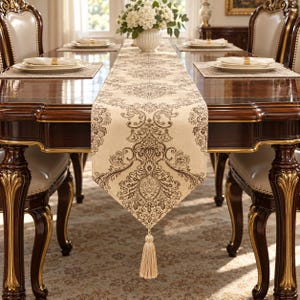 May include: A long, rectangular table runner featuring a cream-colored fabric base with a dark brown, intricate damask pattern. The runner hangs over the edge of a dark wood table and is finished with a single, light beige decorative tassel.