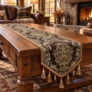 Puede incluir: Un camino de mesa rectangular con una base de tela marrón oscuro y un intrincado patrón de damasco repetitivo en tonos verde oliva y crema. Los bordes están rematados con un ribete trenzado dorado y múltiples borlas colgantes.