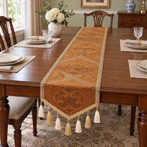 Puede incluir: Un camino de mesa rectangular largo con un rico patrón de damasco marrón dorado y volutas intrincadas. El camino está rematado con un borde trenzado decorativo de color dorado y varias borlas de color crema que cuelgan de los extremos puntiagudos.
