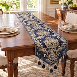 Puede incluir: Un camino de mesa rectangular largo de tela azul marino profundo con un intrincado patrón de damasco repetitivo en tonos dorados y beige. El camino está rematado con un borde decorativo y múltiples borlas colgantes en azul y oro.