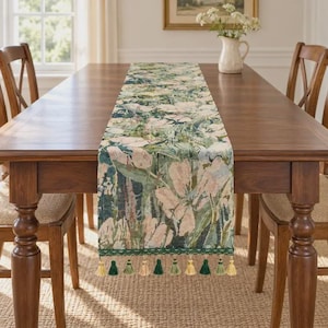 May include: A rectangular table runner featuring a woven floral pattern in shades of green, cream, and soft pink. The runner is finished with a decorative green lace trim and a series of alternating cream and green tassels along the edges.
