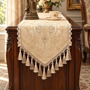 May include: A beige table runner featuring an intricate woven damask pattern, scalloped lace edges, and a pointed end finished with multiple hanging tassels. The fabric displays a textured, monochromatic design with subtle cream and light tan details.