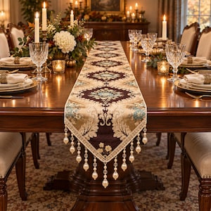 Puede incluir: Un camino de mesa rectangular largo con un patrón de damasco en marrón oscuro y beige con detalles florales en azul claro. El camino está rematado con un borde decorativo y borlas de cuentas colgantes con perlas blancas y cristales.