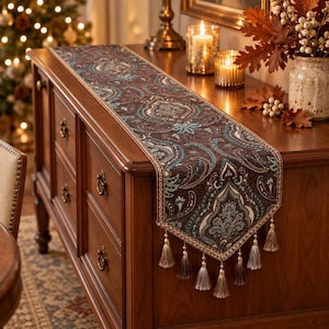 Puede incluir: Un camino de mesa rectangular con una base marrón oscuro y un intrincado patrón de cachemira en tonos azul cerceta, crema y beige. El camino está rematado con un borde decorativo y múltiples borlas colgantes en tonos marrón, crema y gris claro.