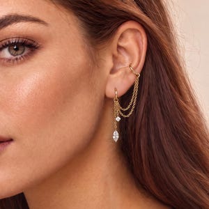 May include: A gold ear cuff connected by two delicate chains to a small hoop earring. The jewelry features a small square-cut clear stone and a larger marquise-cut clear stone dangling from the chains, creating a layered, multi-pierced look.