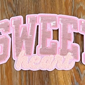 May include: Pink sequined iron-on patch with the text "SWEET" in large letters and "heart" in smaller letters below.