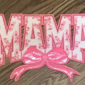May include: Pink felt applique with the word "MAMA" in white letters with a pink floral pattern. A pink sequined bow is centered below the letters.