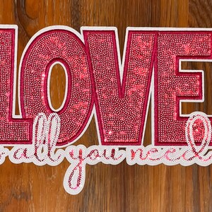 May include: Pink sparkly letters spelling "LOVE is all you need" on a wood background.