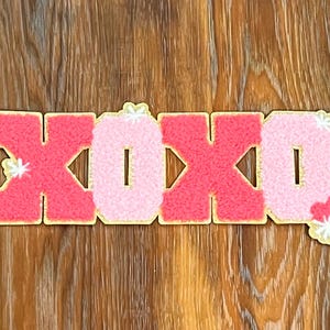 May include: Pink and red felt letters spelling "XOXO" with white glitter accents. The letters are outlined in gold.
