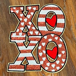 May include: A decorative wooden sign spelling "XO" in red, white, and gold. The "X" is red with white hearts, and the "O"s are red with white stripes and a red heart in the center. The sign is on a wooden surface.