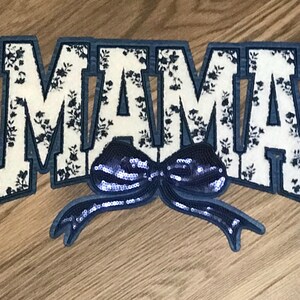 May include: A white fabric patch with the word "MAMA" in blue outlined letters. The letters are covered in a black and white floral pattern. A blue sequined bow is attached to the bottom of the patch.